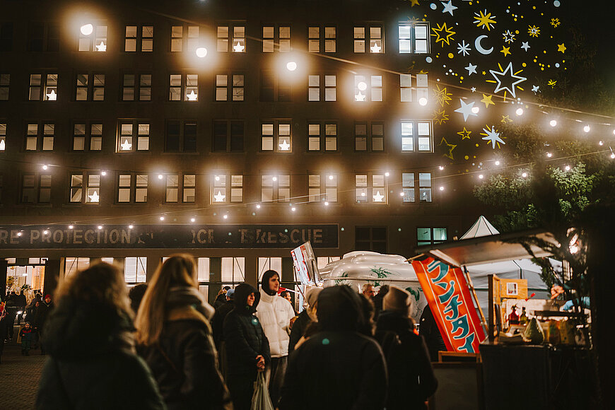 Die weihnachtlich beleuchtete Außenfassade des THF TOWER bei Abend während des Wintermarkt. Menschen stehen Schlange an einem Foodtruck