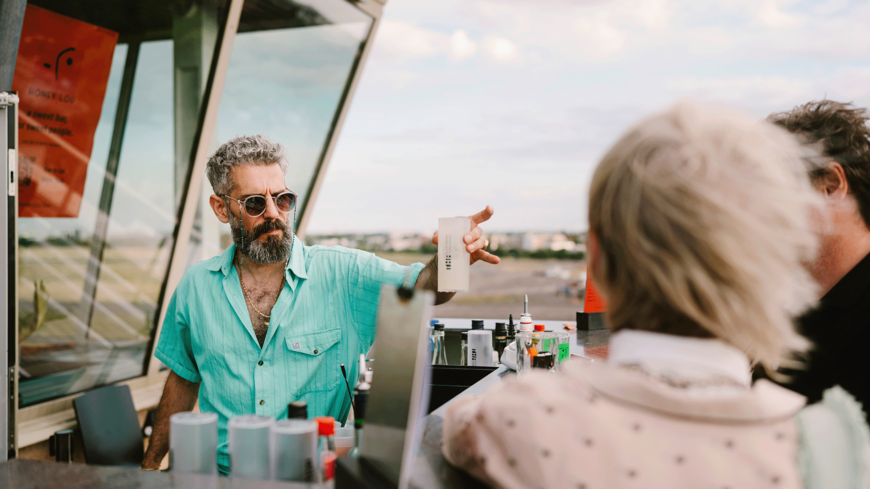 Ein Barkeeper mit grünem Hemd und Sonnenbrille reicht Gästen auf dem THF TOWER einen Drink