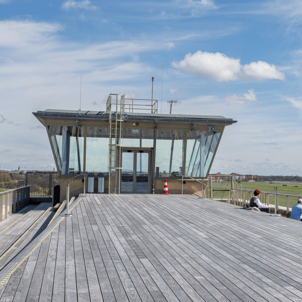 Weite Dachterrasse des THF TOWER mit Blick über das Tempelhofer Feld. Im Vordergrund liegt eine große Holzfläche auf der einzelne Personen sitzen. Die Umgebung ist von Grasflächen umgeben, der Himmel ist leicht bewölkt.