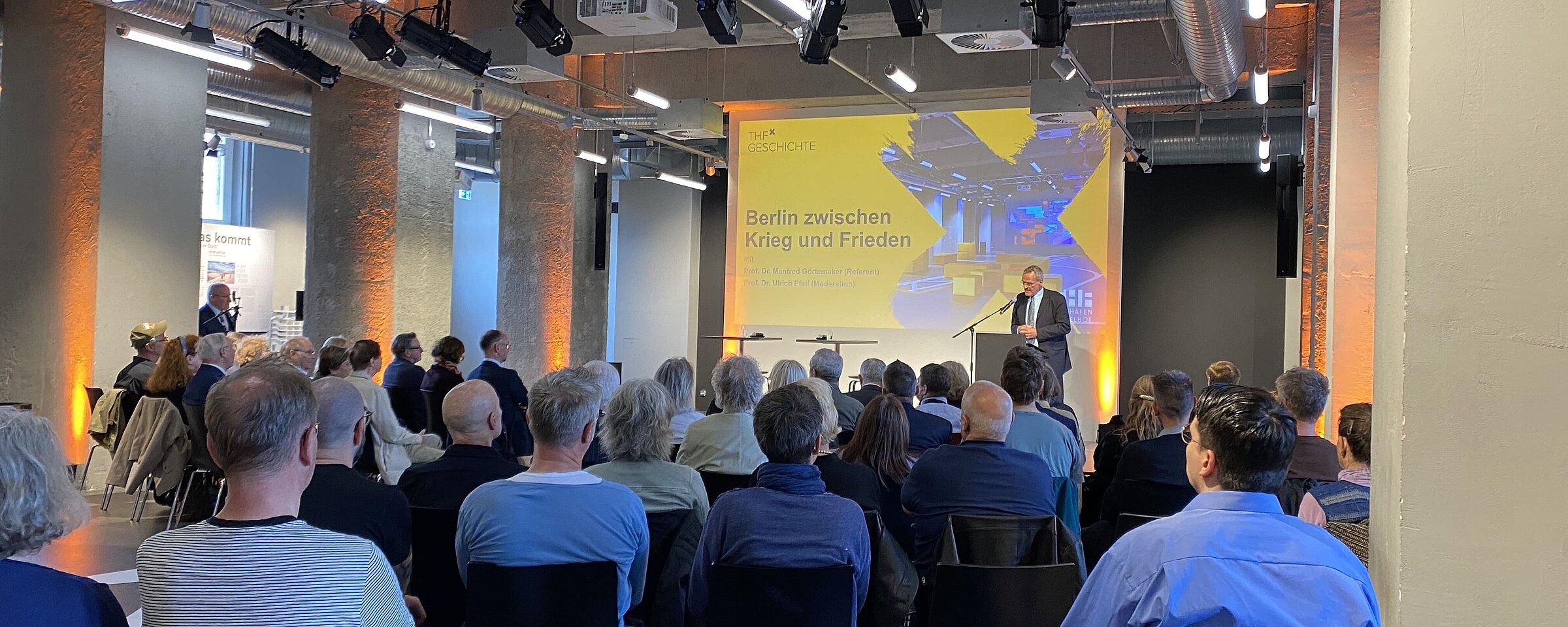 Auf dem Bild sieht man viele Menschen im Besucherzentrum CHECK-IN sitzen. Auf der Bühne trägt jemand etwas vor.