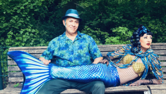 Zwei Personen sitzen auf einer Holzbank im Grünen; eine trägt ein blau gemustertes Hemd und einen blauen Hut, die andere ist als Meerjungfrau mit blauem Fischschwanz und goldenem Oberteil verkleidet, während die erste Person den Schwanz hält.