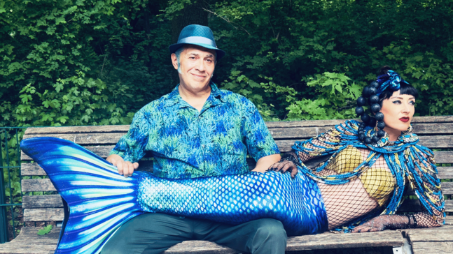 Zwei Personen sitzen auf einer Holzbank im Grünen; eine trägt ein blau gemustertes Hemd und einen blauen Hut, die andere ist als Meerjungfrau mit blauem Fischschwanz und goldenem Oberteil verkleidet, während die erste Person den Schwanz hält.