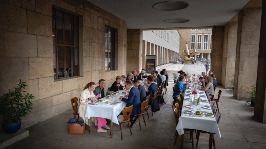 Menschen sitzen an zwei langen Tafeln mit weißen Tischdecken am Flughafen Tempelhof und essen 