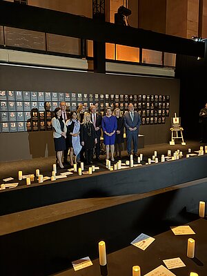 Berliner Senatsmitglieder:innen stehen vor der Gedenkwand in der Nova-Ausstellung am Flughafen Tempelhof. Im Hintergrund hängen Fotos von den Geiseln, im Vordergrund leuchten Kerzen