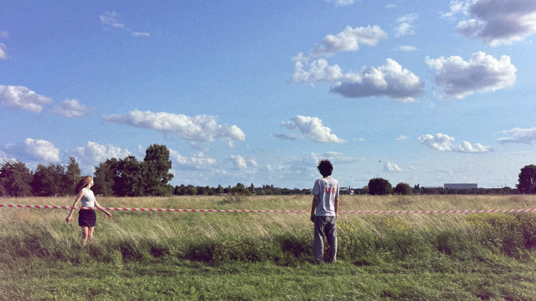 Die Band Calcou stehen in einer weitläufigen Wiese auf dem Tempelhofer Feld unter blauem Himmel. Zwischen ihnen verläuft ein rot‑weißes Absperrband. Links steht eine Person in hellem Oberteil und dunklem Rock, rechts eine Person in hellem Hemd und grauer Hose. Im Hintergrund sind Bäume und vereinzelte Gebäude zu erkennen.