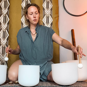 Mona Glass sitzt auf dem Boden vor drei großen weißen Klangschalen und spielt sie mit einem weißen Mallet. Die Szene wirkt ruhig und meditativ; im Hintergrund hängen gemusterte Stoffbahnen und ein rundes Wanddekor.