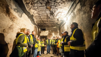 Eine Gruppe von Menschen in gelben Warnwesten steht in einem schmalen, schwach beleuchteten Tunnel mit rohen Betonwänden und beschädigter Decke, aus der Metallstreben hervortreten. Eine einzelne Glühbirne hängt von der Decke und beleuchtet die Szene, die wie eine Führung oder Begehung eines historischen oder technischen Untergrundraums wirkt.
