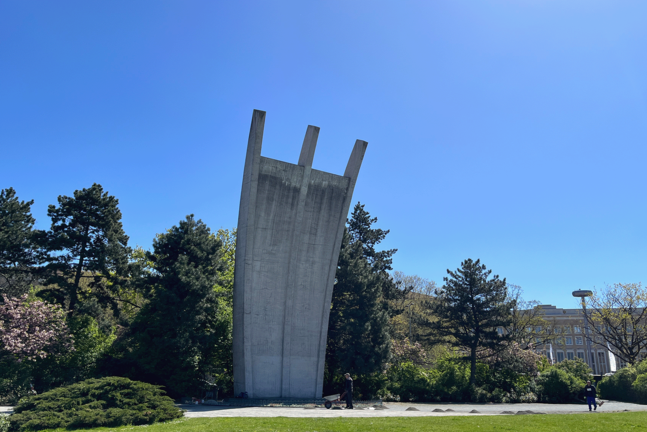 Das Luftbrückendenkmal am Flughafen Tempelhof in Berlin bei strahlend blauem Himmel. Das markante, bogenförmige Betonmonument mit den drei nach oben gerichteten „Krallen“ steht vor dichten grünen Bäumen auf einer gepflegten Rasenfläche.