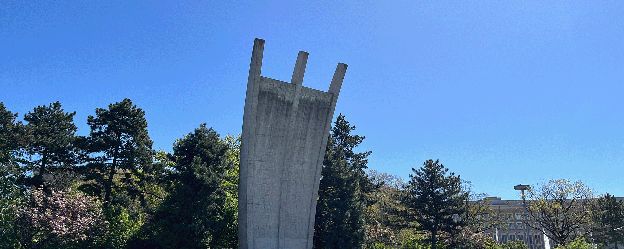 Das Luftbrückendenkmal am Flughafen Tempelhof in Berlin bei strahlend blauem Himmel. Das markante, bogenförmige Betonmonument mit den drei nach oben gerichteten „Krallen“ steht vor dichten grünen Bäumen auf einer gepflegten Rasenfläche.