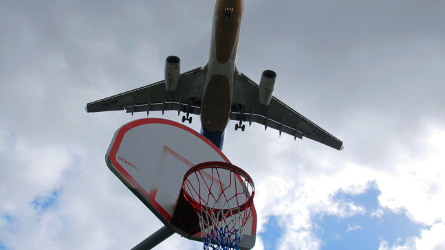 Ein von unten fotografierter Basketballkorb. Im Himmel fliegt ein Flugzeug über den Basketballkorb