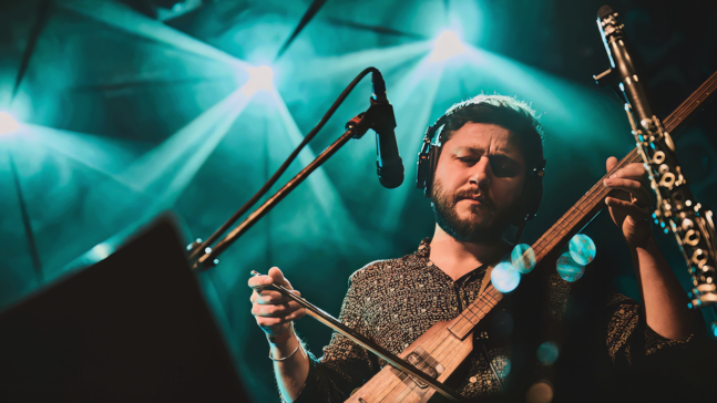 Musiker Kafanar steht auf einer Bühne im türkis‑blauen Licht und spielt ein gestrichenes Saiteninstrument. Er trägt Kopfhörer, steht vor einem Mikrofon und ist in konzentrierter Haltung zu sehen, während kräftige Strahlen der Bühnenbeleuchtung hinter ihm auffächern.