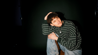 Porträt des Musikers JAS in einem Studio vor dunkelgrünem Hintergrund. Er sitzt auf einem Hocker, trägt ein schwarz‑weiß gestreiftes Langarmshirt, hellblaue Jeans und braune Stiefel. Mit kurz geschnittenem dunklem Haar blickt er ruhig in die Kamera, die Hände locker vor dem Körper verschränkt. Das Licht hebt ihn klar vom gedimmten Hintergrund ab.