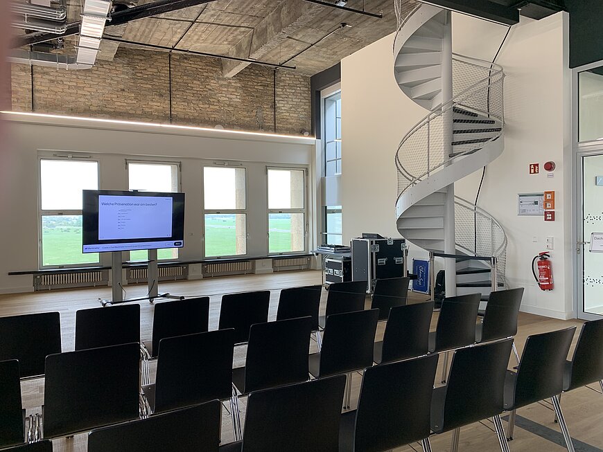 Raum im THF TOWER mit Reihen schwarzer Stühle, großem Display in der Mitte, einer hellen Fensterfront und einer weißen Wendeltreppe an der Seite.