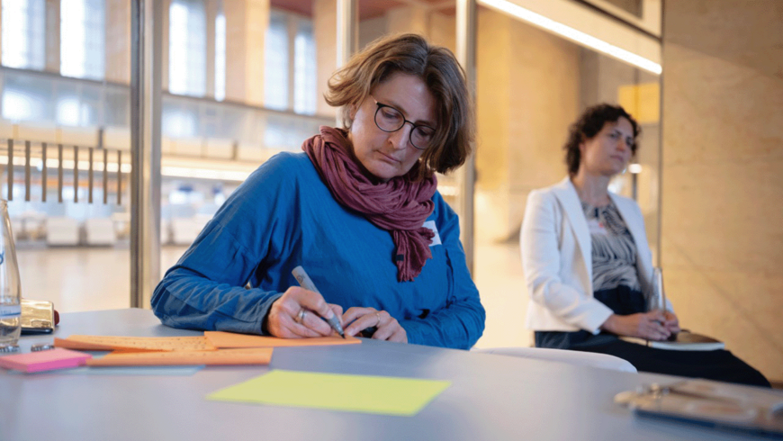 Zwei Frauen an einem Schreibtisch mit bunten Karten. Eine schreibt, die andere hört zu. Im Hintergrund die Haupthalle des Flughafen Tempelhof