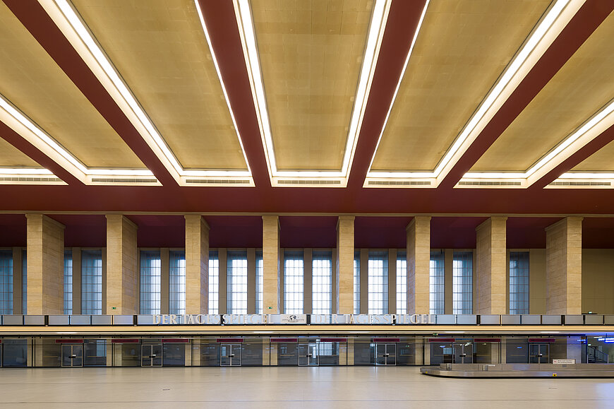Querblick in die Haupthalle am Flughafen Tempelhof mit symmetrischen Säulen am Ende des Bildes und Lampen an den hohen Decken