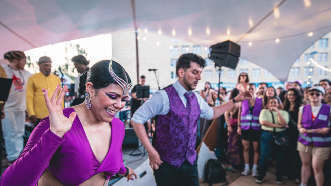 Eine lebhafte Szene beim Berlin en Salsa Festival am THF TOWER unter einem großen, überdachten Außenbereich mit Lichterketten. Im Vordergrund tanzen zwei Personen in auffälligen lilafarbenen Outfits, während im Hintergrund eine Gruppe von Menschen zuschaut und lächelt