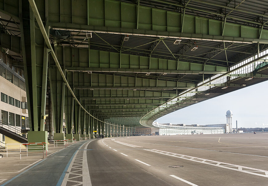 Überdachtes Vorfeld am Flughafen Tempelhof mit grüner Stahlkonstruktion. Im Hintergrund ragt der Radarturm in den Himmel.