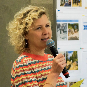 Ela Kagel spricht in ein Mikrofon, trägt einen farbig gemusterten Pullover und steht vor einer Präsentationswand mit ausgedruckten Fotos und Texttafeln.