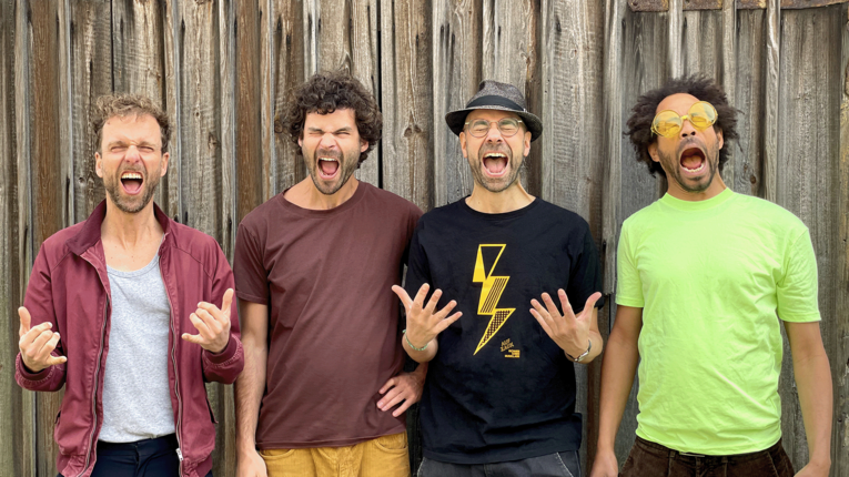 Die Band Richards Kindermusikladen  vor einer Holzverkleidung und machen übertriebene, humorvolle Grimassen und Gesten. Sie tragen farblich unterschiedliche Oberteile – rot, bordeaux, schwarz und leuchtend grün – wobei die Person rechts einen Gegenstand vor einem Auge hält.