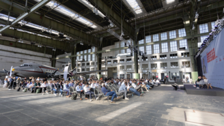 Blick in den Hangar am Flughafen Tempelhof während der ICNC: Viele Menschen sitzen auf Stühlen und schauen auf eine Präsentation. Im Hintergrund ein Flugzeug