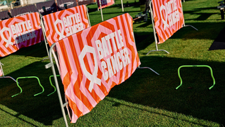 Absperrgitter auf einer Wiese mit orange‑weißen Bannern der Aktion „Battle Cancer“; davor stehen kleine grüne Trainingshürden für sportliche Aktivitäten.