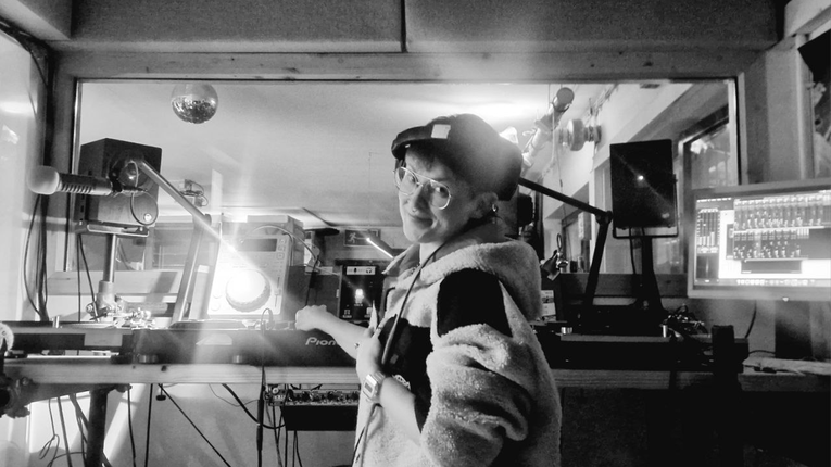 Person mit Brille und Cap steht in einem Tonstudio vor Mischpult, Mikrofonarm, Lautsprechern und einem Computerbildschirm mit Audioprogramm; im Vordergrund ein beleuchteter Plattenspieler von Pioneer, umgeben von Kabeln und Studioequipment.