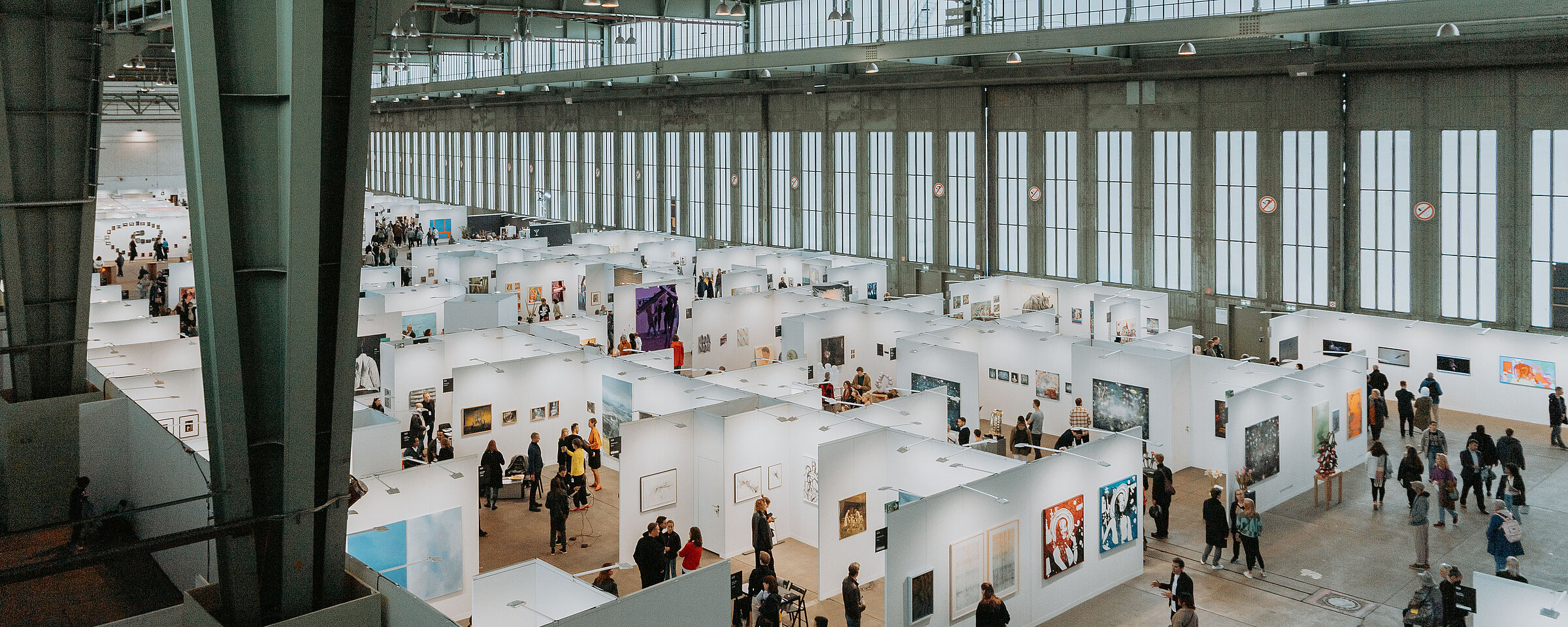 Ausstellung in einem Hangar am Flughafen Tempelhof mit vielen Besuchern, die Kunstwerke an weißen Ausstellungsständen betrachten.