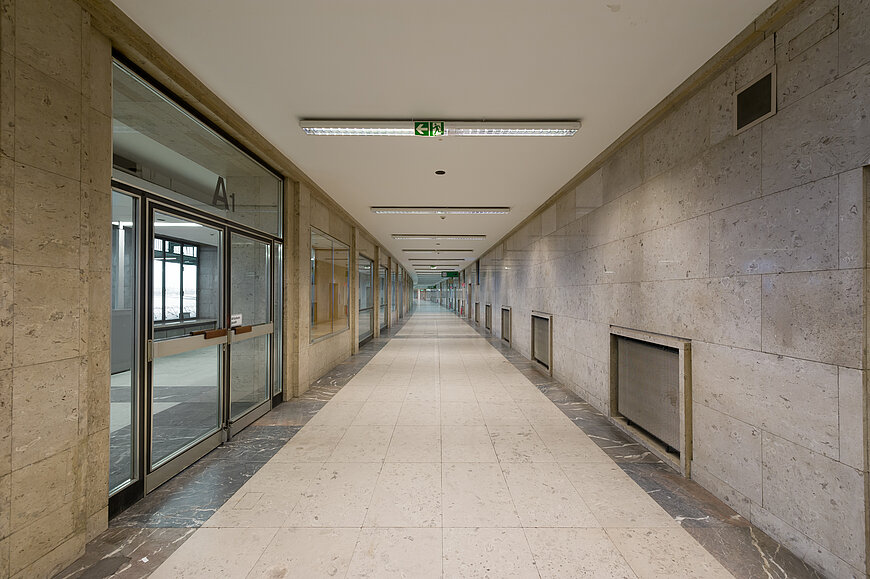 Ein langer, symmetrischer Transitgang im Flughafen Tempelhof. Der Boden besteht aus hellen, beigefarbenen Fliesen mit einem dunkleren Marmorstreifen entlang der Ränder. Die Wände sind mit großen, strukturierten Natursteinplatten verkleidet. Auf der linken Seite befindet sich eine durchgehende Glasfront mit mehreren Fenstern und einer Doppeltür im Vordergrund (beschriftet mit "A1"), die in einen helleren Bereich führt. An der Decke hängen mehrere parallel verlaufende Leuchtstoffröhren. In der Mitte der Decke, über dem Flur, hängt ein beleuchtetes grünes Piktogramm, das den Notausgang in Pfeilrichtung anzeigt. Der Flur verjüngt sich stark in die Ferne, was einen tiefen Fluchtpunkt erzeugt. An der rechten Wand sind mehrere tiefe, vergitterte Aussparungen oder Lüftungsschächte erkennbar.