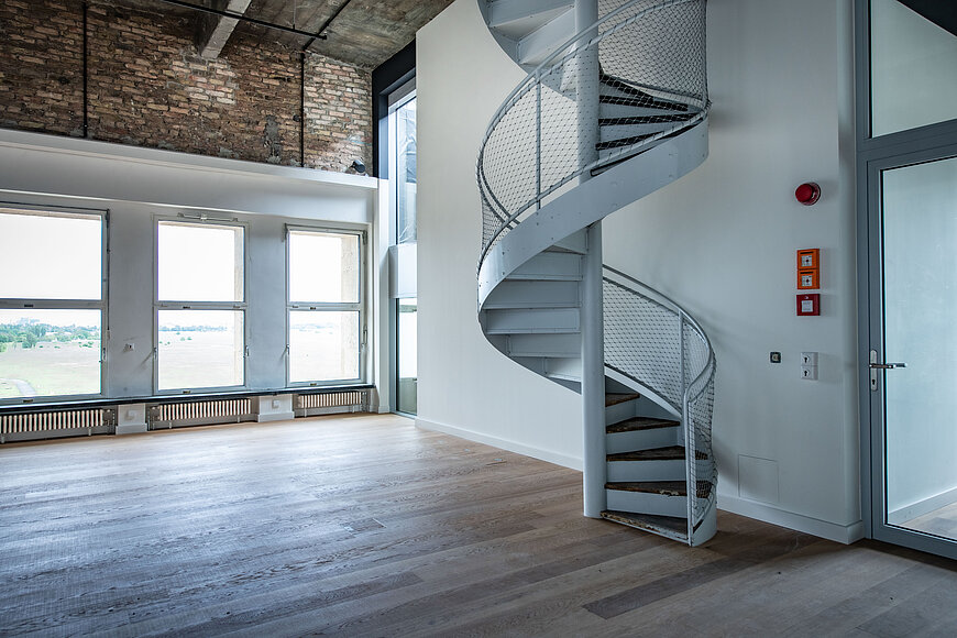 Eventlocation in der 6. Etage des THF TOWER mit hellen Holzdielen und weißen Wänden. Der Raum verfügt über drei große Fenster, die viel Licht hereinlassen und einen Blick nach draußen bieten.  Ein weiße, metallene Wendeltreppe mit Maschendrahtgeländer ist prominent im vorderen rechten Bereich platziert und führt zu einer oberen Ebene.