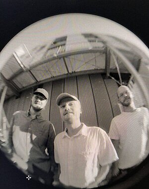 Drei Personen stehen eng beieinander im Freien vor einer hellen Gebäudewand; die Aufnahme ist mit einem Fisheye‑Objektiv gemacht, wodurch die Bildränder stark gekrümmt wirken.