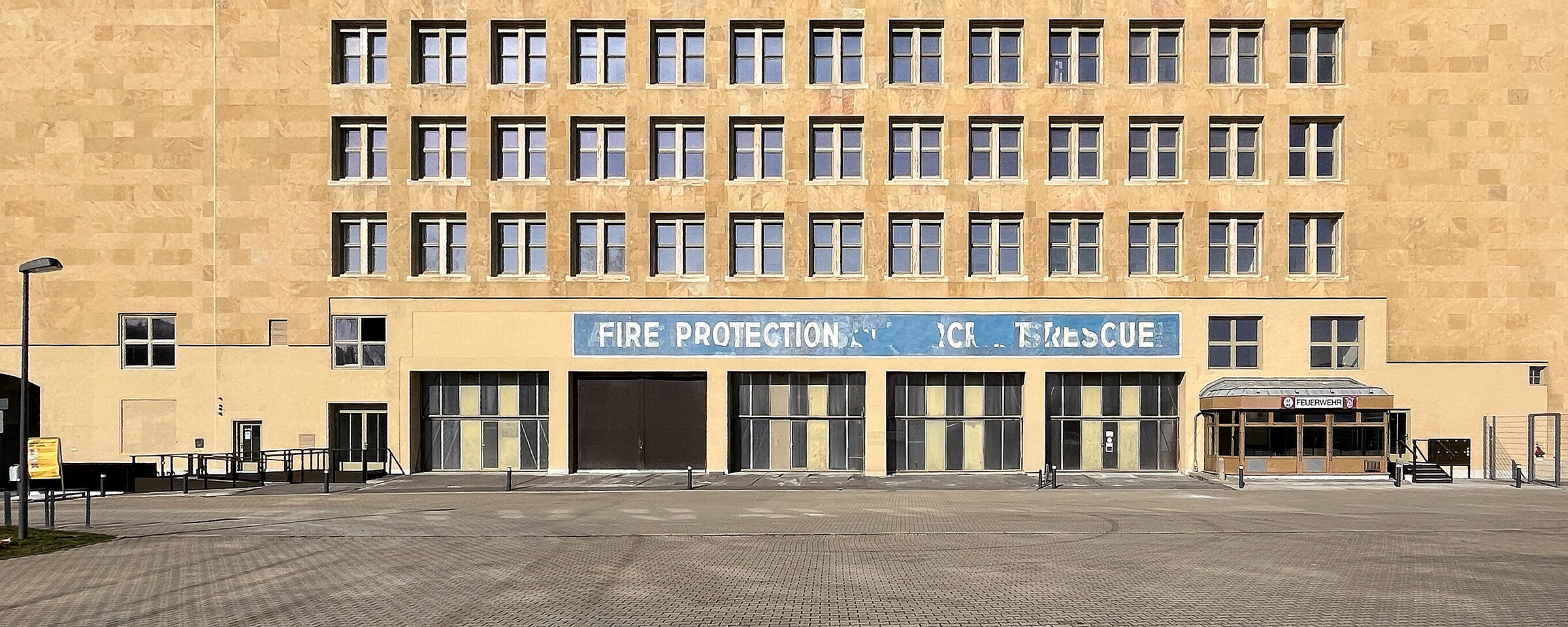 Die Fassade der Alten Feuerwache mit ihrem hellen Steinbau und den gleichmäßig angeordneten Fenstern. Über dem Eingang steht der Schriftzug "Fire Protection". Vor dem Gebäude herrscht klare Sicht unter blauem Himmel.