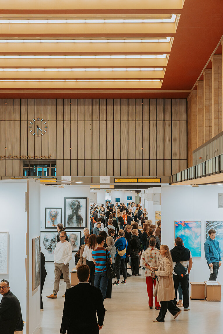 Besuchende laufen durch die Ausstellung Paper Positions in der Haupthalle des Flughafen Tempelhof. Im Hintergrund der ikonische "Restaurant"-Schriftzug und eine Uhr