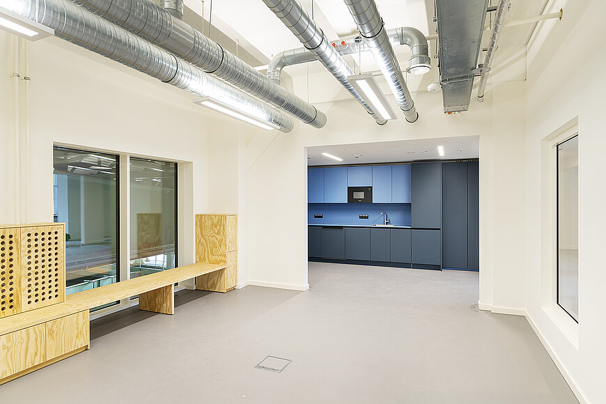 Innenansicht eines modernen, hellen Büroraums im Kopfbau West des Flughafens Tempelhof. Der Raum verfügt über eine große, freie Fläche mit einem hellgrauen Boden und hellbeigen Wänden. Auffällig ist die offene Deckeninstallation mit sichtbaren, großen Lüftungsrohren aus Metall und länglichen Deckenleuchten. Im hinteren Bereich befindet sich eine Küchenzeile mit blauen und dunkelgrauen Schränken. Links im Vordergrund ist eine maßgefertigte Sitzgelegenheit aus hellem, naturbelassenem Sperrholz mit integriertem Stauraum und einer langen Holzbank zu sehen