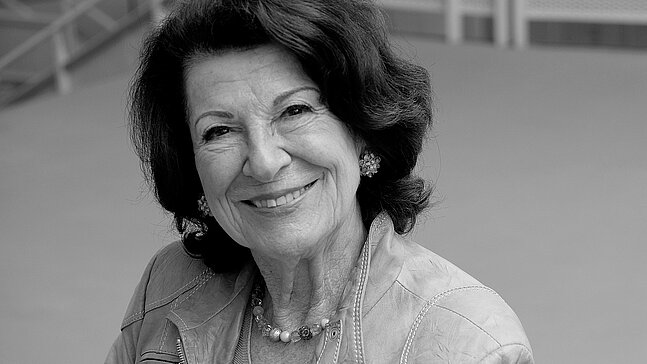 Schwarz-Weiß-Porträt von Irene Bindel mit dunklem, schulterlangem Haar. Die Person blickt in die Kamera und trägt eine helle Jacke, Ohrschmuck und eine Perlenkette. Im Hintergrund sind unscharfe architektonische Elemente und ein Geländer zu sehen.“