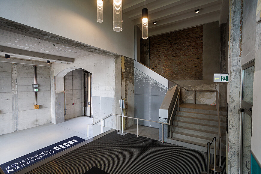 Innenraum des modernen, industriell gestalteten Eingangsbereichs des THF TOWER mit rohen Betonwänden, einer Treppe mit schwarz-weißem grafischem Muster und einem ebenfalls grafisch gestalteten Boden. Mehrere Hängeleuchten hängen von der Decke