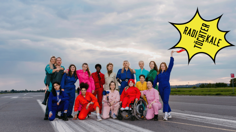 Gruppe von 14 Personen in verschiedenfarbigen Overalls auf dem Tempelhofer Feld. Eine Person sitzt im Rollstuhl, mehrere Personen halten Megafone. Im rechten oberen Bildbereich befindet sich ein gelber Stern mit der Aufschrift ‚Radikale Töchter‘ vor bewölktem Himmel.