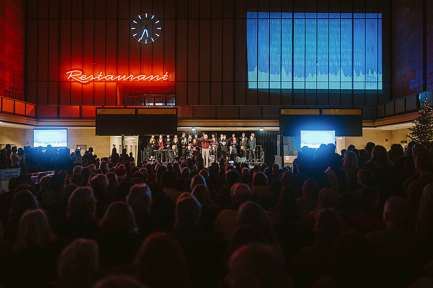 Haupthalle am Flughafen Tempelhof ist gefüllt mit Menschen. Im Hintergrund leuchtet der rote Schriftzug "Restaurant" und an der Wand ist ein blaues Bild mit einem Beamer projiziert. 