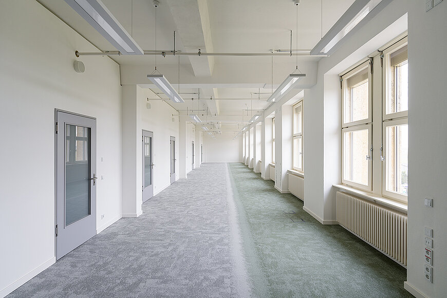 Langer, heller Bürogang im sanierten Kopfbau West des Flughafen Tempelhof mit grau-grünem Teppichboden, vielen Säulen und einer Reihe hoher Fenster mit Heizkörpern.