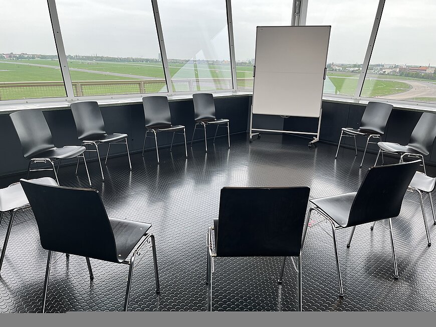 Raum im Fluglotsentower, eingerichtet für eine Konferenz oder Präsentation. Der Raum verfügt über Reihen schwarzer Stühle auf einem hellen Holzboden und wird von weißen Ausstellungsmodulen strukturiert, die Infotafeln und hängende Monitore mit grafischen und thematischen Inhalten zeigen. Die Decke ist schwarz und industriell gehalten, und rechts befindet sich eine Wand mit hohen Fenstern.