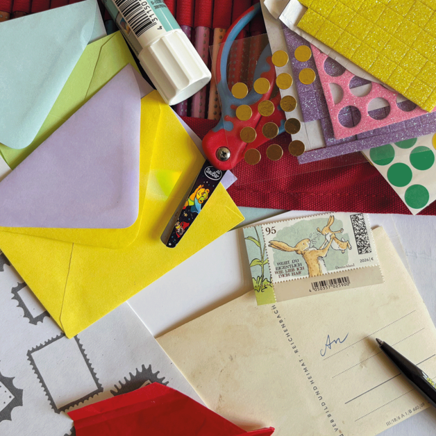Bunte Bastelmaterialien liegen auf einer Fläche verteilt: farbige Briefumschläge, Stickerbögen mit Punkten und Glitzerstrukturen, eine Kinderschere mit Motiv, ein Klebestift, ein Stift, mehrere Briefmarken sowie ein Blatt mit vorgedruckten Briefmarkenrahmen und deutschem Text.