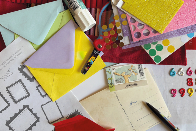 Bunte Bastelmaterialien liegen auf einer Fläche verteilt: farbige Briefumschläge, Stickerbögen mit Punkten und Glitzerstrukturen, eine Kinderschere mit Motiv, ein Klebestift, ein Stift, mehrere Briefmarken sowie ein Blatt mit vorgedruckten Briefmarkenrahmen und deutschem Text.