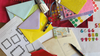 Bunte Bastelmaterialien liegen auf einer Fläche verteilt: farbige Briefumschläge, Stickerbögen mit Punkten und Glitzerstrukturen, eine Kinderschere mit Motiv, ein Klebestift, ein Stift, mehrere Briefmarken sowie ein Blatt mit vorgedruckten Briefmarkenrahmen und deutschem Text.