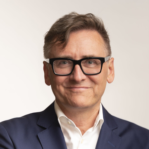 Heinz Jirout in einem professionellen Porträt: Er trägt eine dunkle Brille, ein weißes Hemd und ein dunkelblaues Sakko, blickt direkt in die Kamera vor hellem Hintergrund.