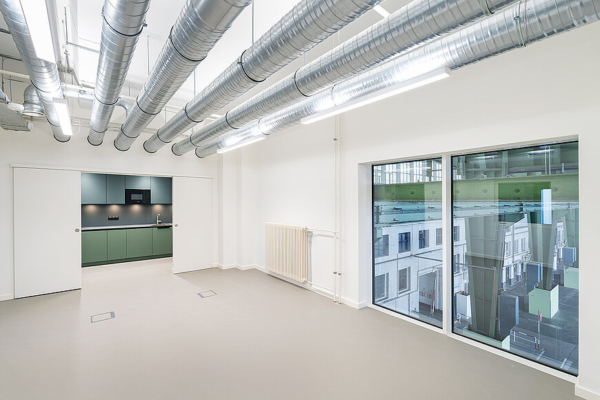 Innenansicht eines modernen, sanierten Büros am Flughafen Tempelhof (Kopfbau West). Der helle Hauptraum ist leer und verfügt über große Fenster, die viel Licht hereinlassen. Die Decke ist offen gestaltet mit vielen sichtbaren, glänzenden Lüftungsrohren aus Metall und länglichen Leuchten. Durch eine breite Schiebetür im Hintergrund ist ein separater Küchenbereich zu sehen, der durch die Farbwahl der Einbaumöbel einen starken Kontrast bietet: dunkelgrüne Unterschränke und dunkelblaue Oberschränke.