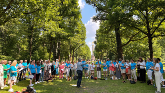 Eine große Gruppe von Menschen steht in einem grünen Park und singt gemeinsam, vor ihnen ein Dirigent