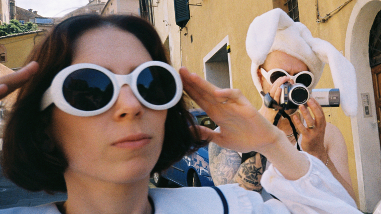 Baby Smith posieren in einer sonnigen Gasse. Im Vordergrund steht eine Person mit kurzen Haaren, weißer runder Sonnenbrille und hellen Kleidungsstücken, die beide Hände an den Kopf legt. Dahinter filmt die zweite Person mit einer kleinen Videokamera und trägt eine weiße Mütze mit langen Hasenohren sowie ebenfalls weiße runde Sonnenbrillen. Im Hintergrund sind gelbe Hausfassaden, Fensterläden und ein geparktes Auto zu sehen.