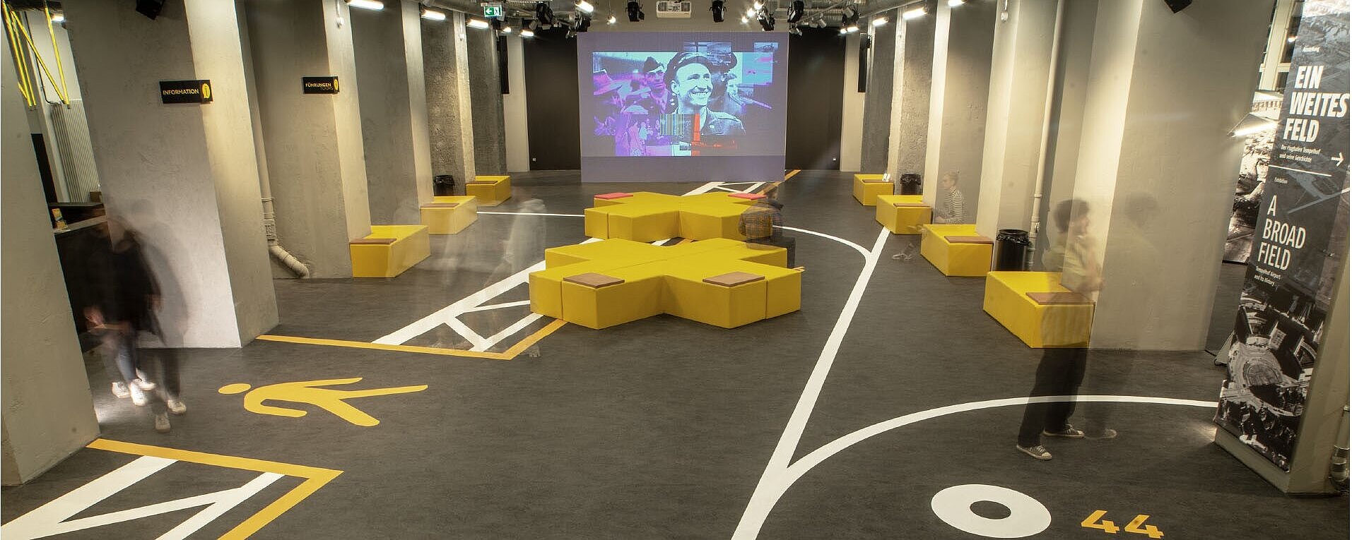 Blick in das Besucherzentrum CHECK-IN mit Videoleinwand und gelben Sitzhockern in der Mitte, schemenhaft sind Besucher und Besucherinnen zu sehen