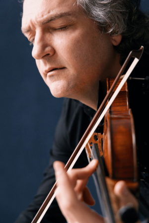 Nahaufnahme des Musikers Ali Moraly, der eine Geige unter dem Kinn hält und mit geschlossenen Augen spielt. Der warme Holzton des Instruments hebt sich klar vom dunklen Hintergrund ab, der Moment wirkt konzentriert und ausdrucksstark.