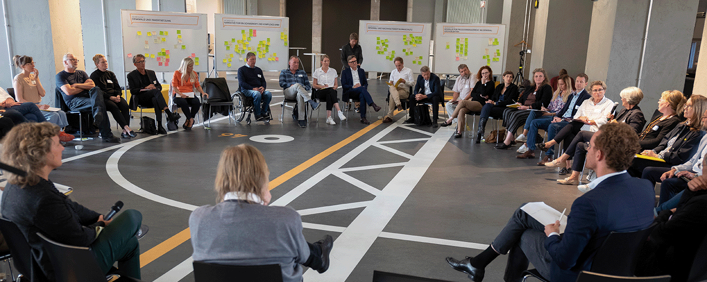 Großer Stuhlkreis im Besucherzentrum mit Menschen, die einr Person zu hören. Im Hintergrund Stellwände mit bunten Post-its