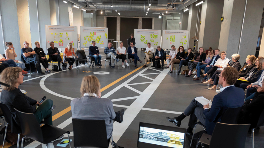 Großer Stuhlkreis im Besucherzentrum mit Menschen, die einr Person zu hören. Im Hintergrund Stellwände mit bunten Post-its